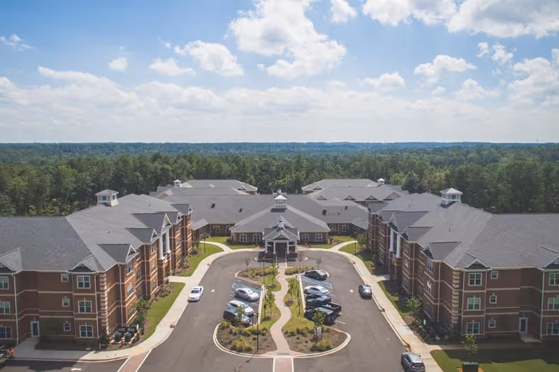 Aerial view of a large brick senior living complex with a circular driveway, parked cars, and surrounding trees under a partly cloudy sky.