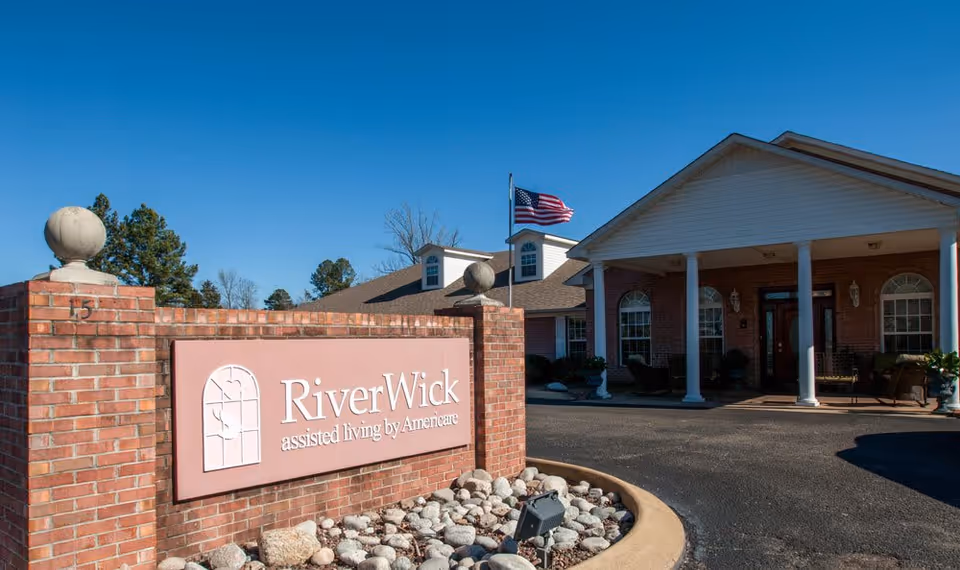 Brick entrance sign reading "RiverWick assisted living by Americare" beside a driveway leading to a single-story brick building with white columns and an American flag.