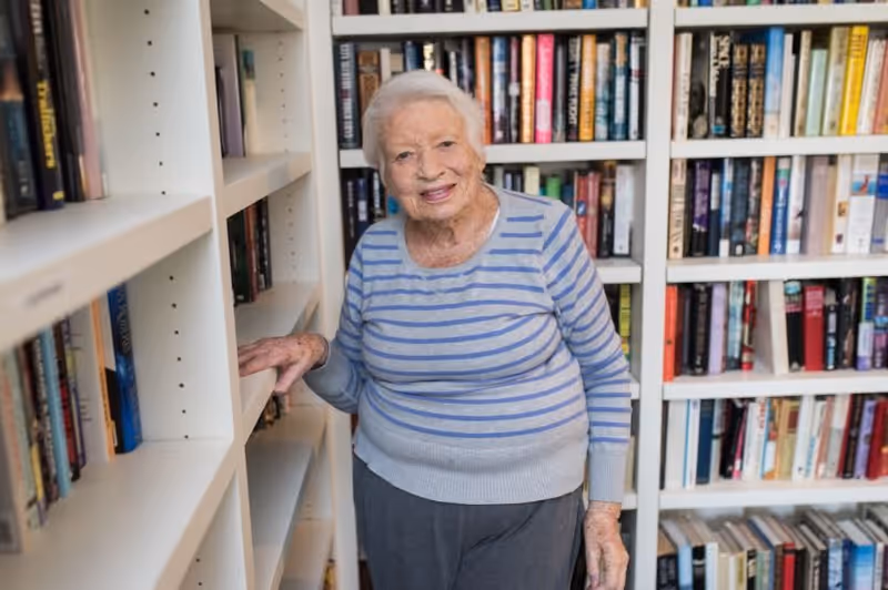 An elderly woman with short white hair wearing a gray and blue striped sweater and gray pants stands smiling in front of white bookshelves filled with books in a library or reading room.