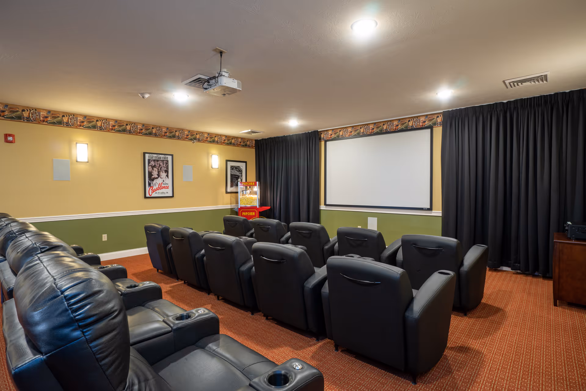 A small movie theater room with two rows of black leather recliner chairs facing a large white projection screen. There is a popcorn machine in the corner, movie posters on the wall, and black curtains framing the screen. The room has a patterned carpet and soft lighting from ceiling fixtures and wall sconces.