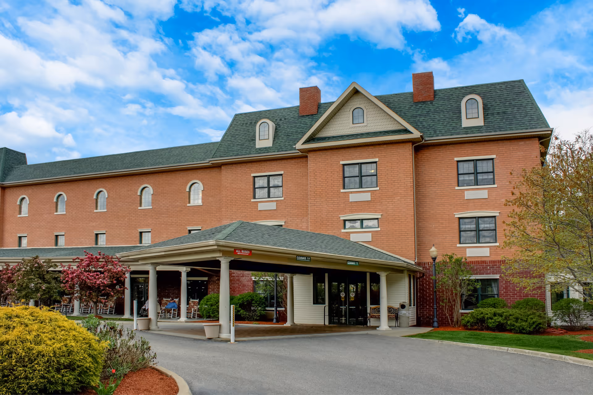 Exterior view of a multi-story brick building with a covered entrance driveway. The building has a green roof with dormer windows and several chimneys. There are landscaped bushes and trees around the entrance area, and a clear blue sky with some clouds above.