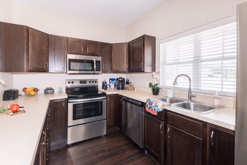 A modern kitchen with dark wooden cabinets, stainless steel appliances including a stove, microwave, dishwasher, and refrigerator. The kitchen has a double sink under a window with white blinds, and various kitchen items like a toaster, fruit bowl, coffee maker, and a colorful towel hanging on the sink.