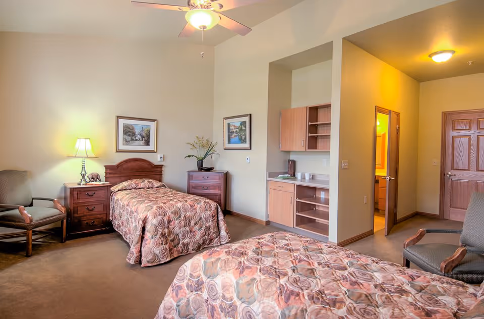 A well-lit bedroom in Countryside Manor featuring two twin beds with patterned bedspreads, a wooden nightstand with a lamp, two armchairs, a wooden dresser with a vase of flowers, framed artwork on the walls, a ceiling fan with light, and an open doorway leading to a bathroom.