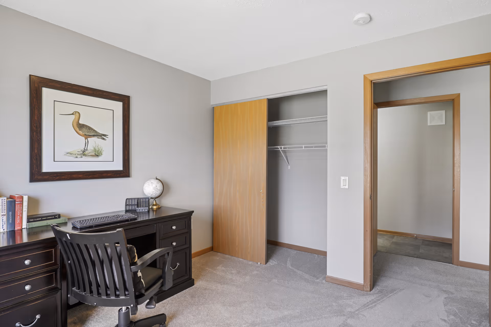 Bedroom with a dark wood desk and chair, an open sliding-wood closet, and framed bird artwork on a neutral-colored wall.