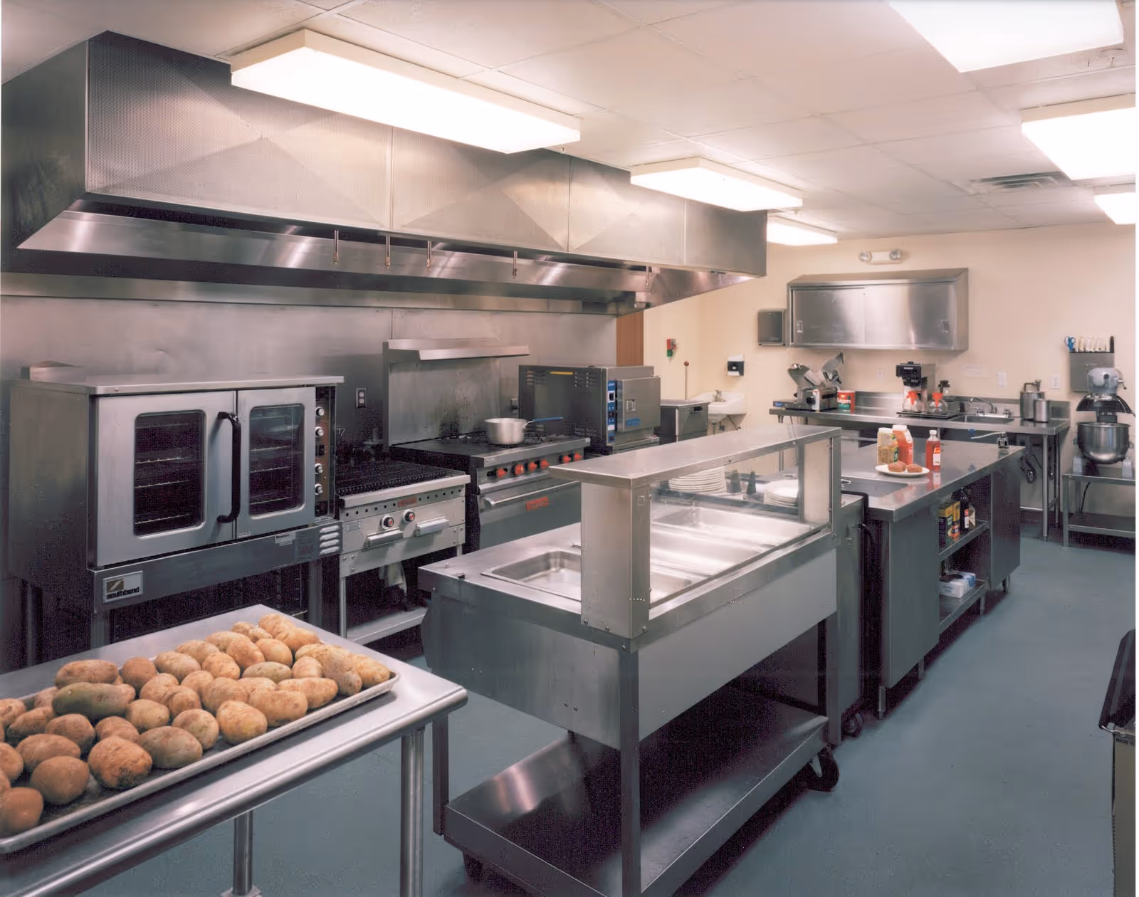 A commercial kitchen with stainless steel appliances and workstations. There is a large oven, stove, and ventilation hood along the back wall. A tray of potatoes is on a table in the foreground. Various kitchen equipment and condiments are visible on the counters.