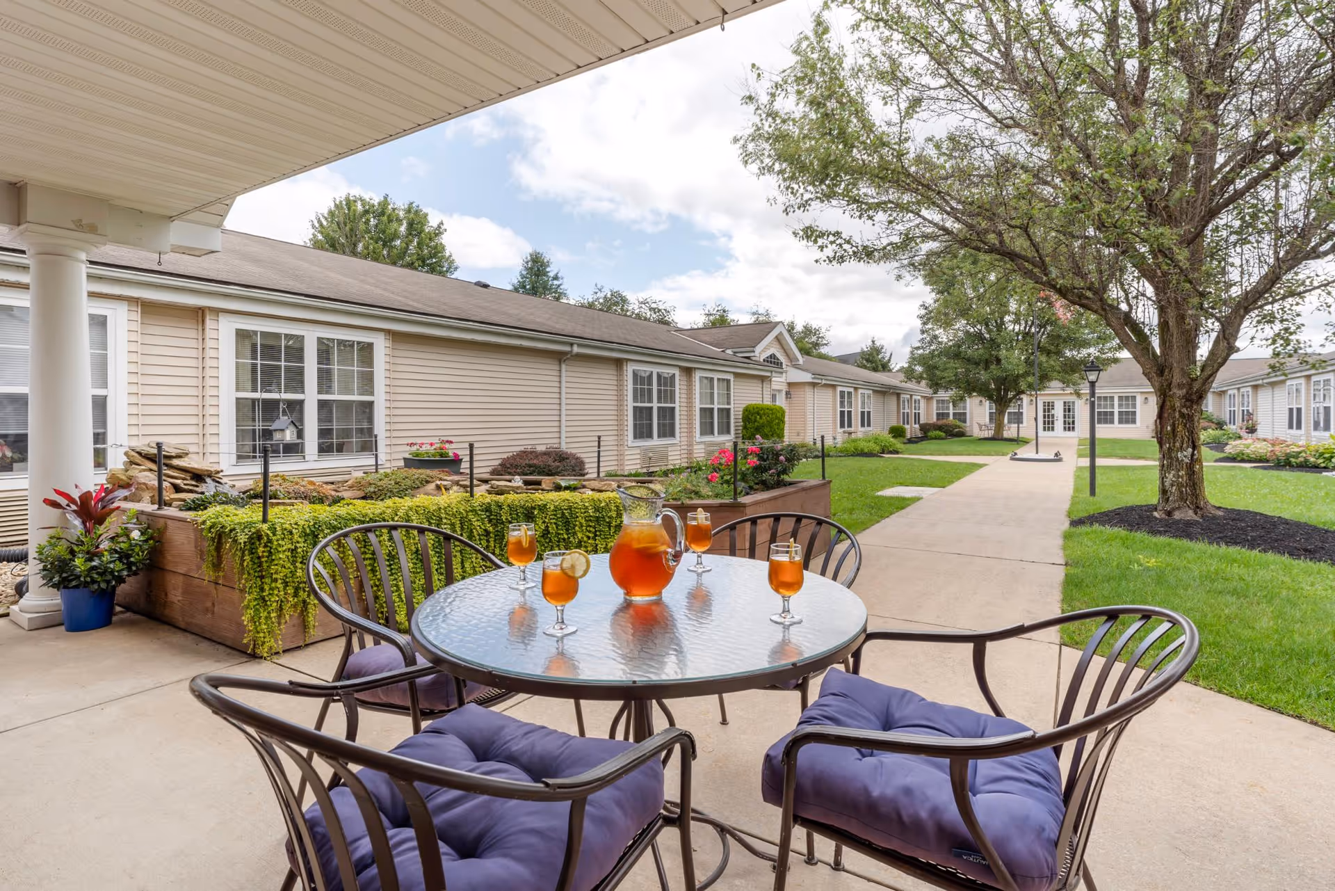 Outdoor patio area at The Addison of Moorehead Place with a round glass table and four metal chairs with purple cushions. On the table is a pitcher and four glasses filled with iced tea. The patio overlooks a walkway, green lawn, trees, and beige single-story buildings under a partly cloudy sky.