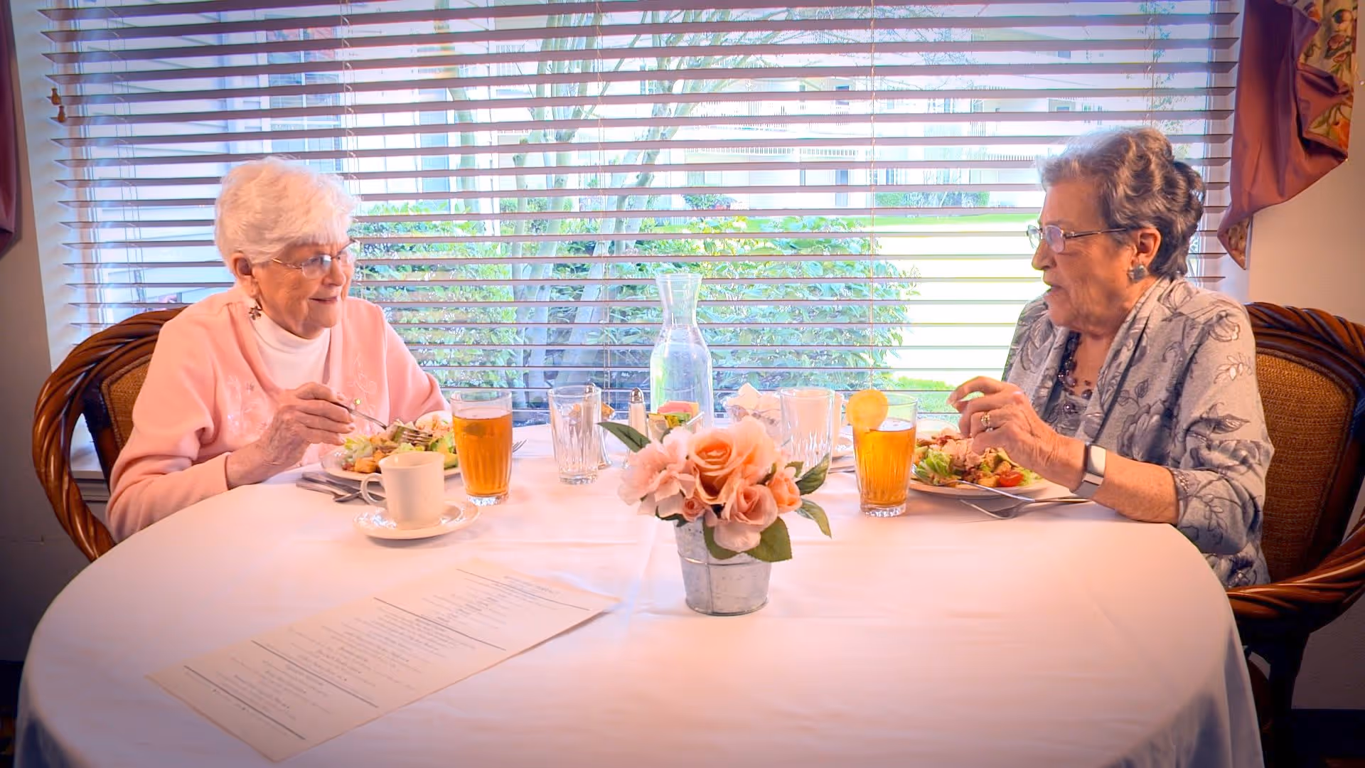 Two elderly women sitting at a round dining table with a white tablecloth, enjoying a meal together. The table has a small flower arrangement in the center, glasses of iced tea with lemon, a coffee cup, and a menu. Behind them is a large window with horizontal blinds, showing greenery outside.