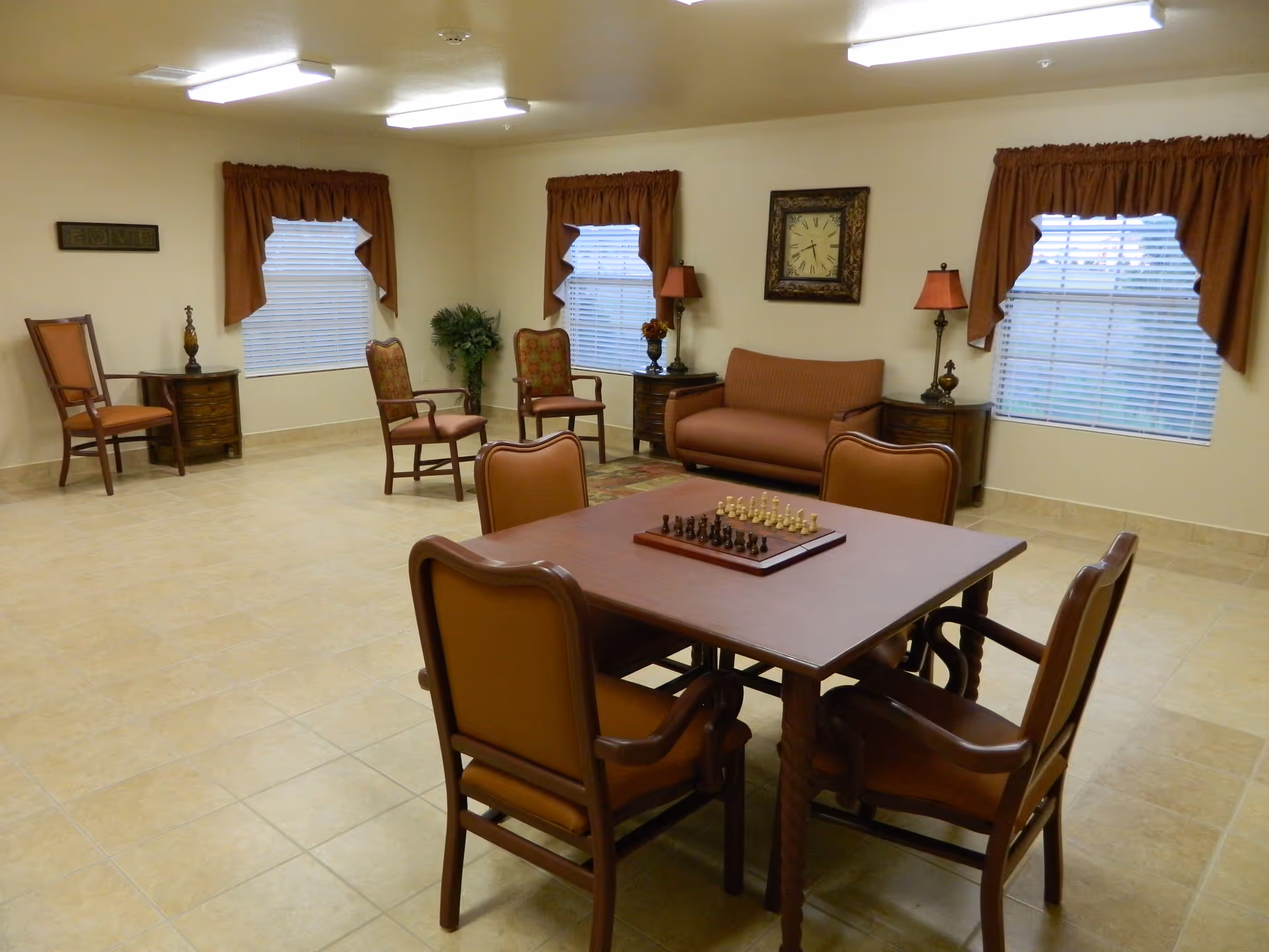 A well-lit common room with a square wooden table and four chairs in the center, featuring a chessboard on the table. The room has beige tiled flooring, cream-colored walls, three windows with brown curtains, a brown sofa, several wooden chairs, side tables with lamps, a wall clock, and a potted plant in the corner.