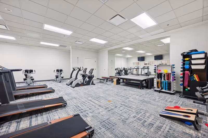 A spacious fitness room with several treadmills, stationary bikes, and exercise benches. The room has a large mirror on one wall, various exercise equipment including weights and resistance bands, and a cornhole game set on the carpeted floor. The ceiling has recessed lighting and the walls are painted white.