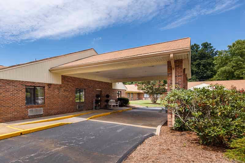 Covered driveway and entrance canopy of a single-story brick senior living facility with shrubs and a blue sky.