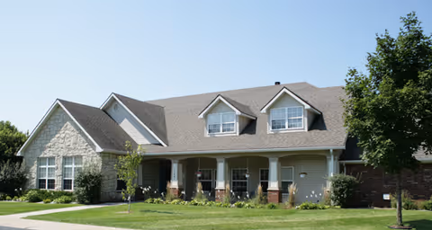 A single-story residential building with a combination of stone and brick exterior, featuring a gable roof with two dormer windows. The front porch has white columns and there is a well-maintained lawn with small trees and shrubs in front of the building under a clear blue sky.