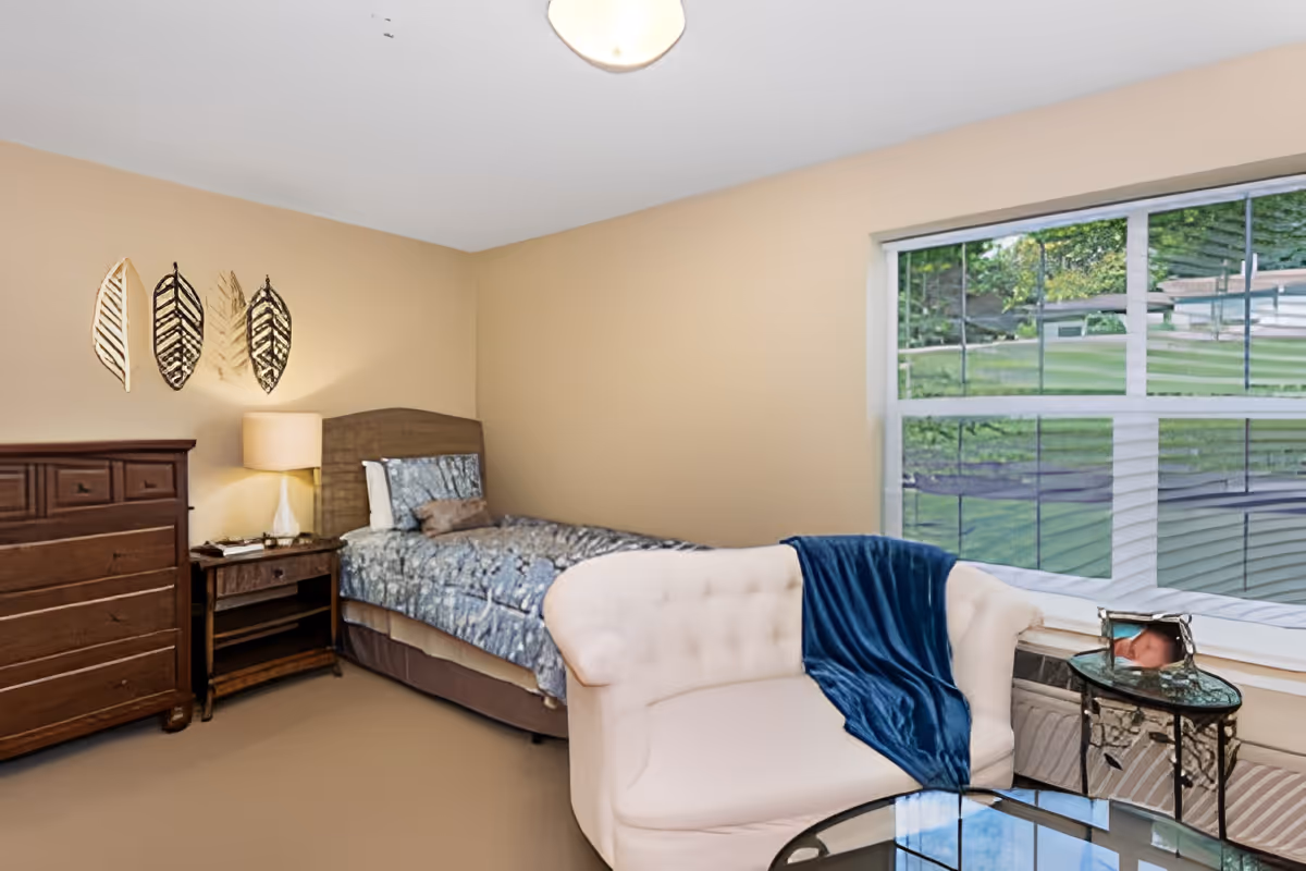 A bedroom with beige walls and carpet featuring a single bed with patterned bedding, a wooden dresser, a wooden nightstand with a lamp, a white tufted loveseat with a blue throw blanket, a glass-top side table, and a large window showing a green outdoor area.