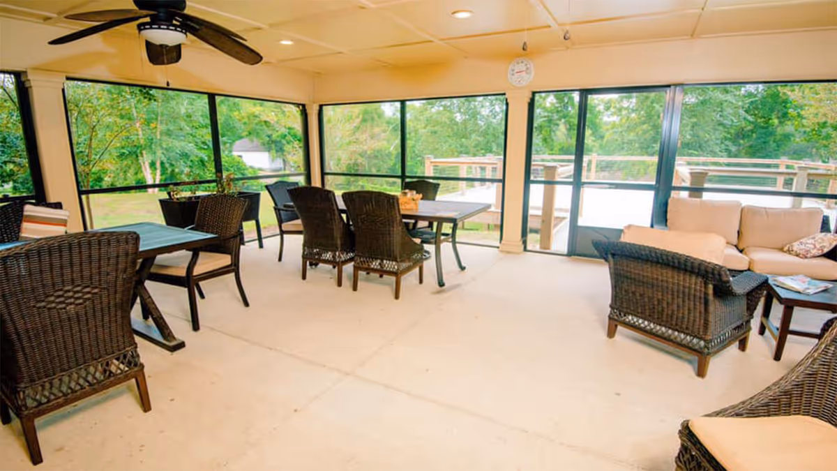 A bright and spacious indoor seating area with large windows offering views of green trees outside. The room features several dark wicker chairs with cushions, two tables, a beige cushioned sofa, a ceiling fan, and a sliding glass door leading to an outdoor deck.