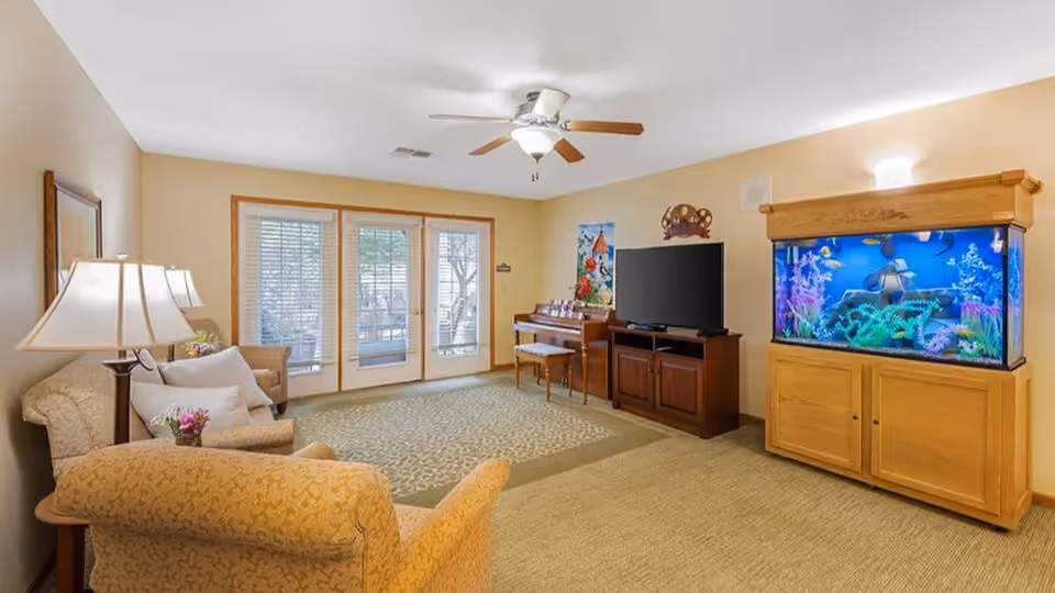 A cozy living room with beige walls and carpet, featuring a patterned armchair and sofa with white pillows, a wooden side table with a lamp and flowers, a large flat-screen TV on a wooden cabinet, a piano with a bench, and a large aquarium with colorful fish and decorations. French doors with blinds lead outside, and a ceiling fan with a light is mounted on the ceiling.