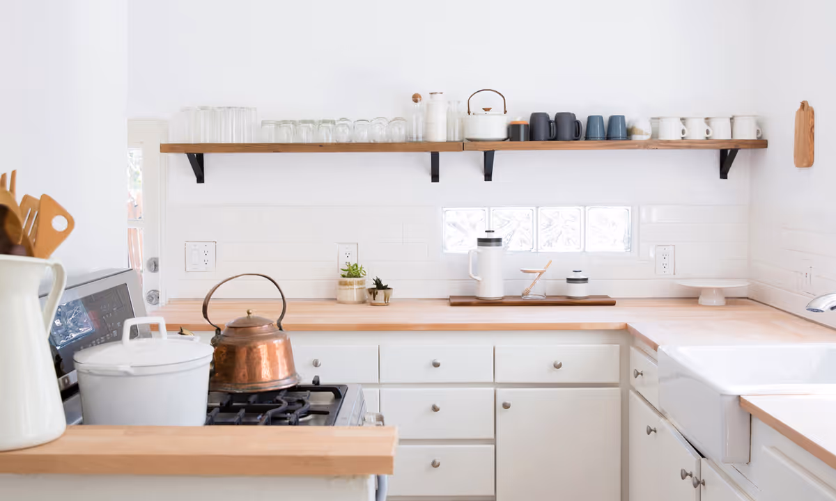 Bright and clean kitchen with white cabinets and wooden countertops. A copper kettle and white pot sit on a gas stove. Above the counter, a wooden shelf holds various glassware, mugs, and a teapot. The backsplash is white tile with a small glass block window. Kitchen utensils are stored in a white pitcher on the counter.
