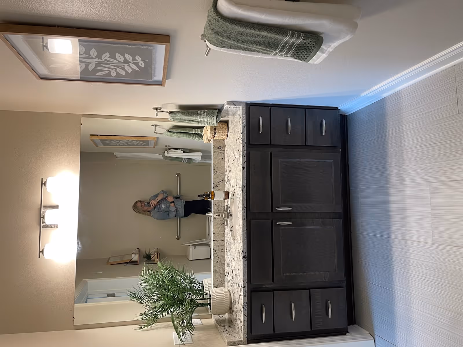 A bathroom with a large mirror above a granite countertop vanity with dark wood cabinets. The countertop has a potted plant, a soap dispenser, and a small basket. Green and white towels hang on the wall to the right, and a framed botanical print is also visible. A person taking a photo is reflected in the mirror, along with a toilet and grab bar in the background.