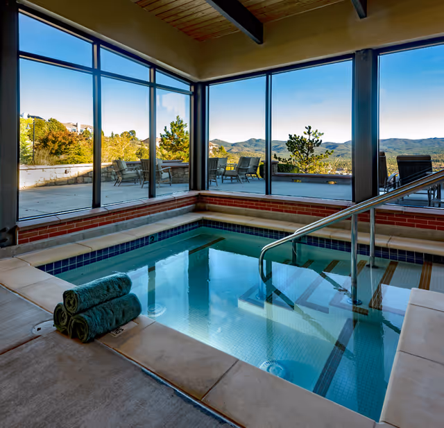 Indoor hot tub with clear water and metal handrails, surrounded by large windows offering a scenic view of trees, patio furniture, and distant mountains under a clear blue sky. Three rolled green towels are placed on the edge of the hot tub.