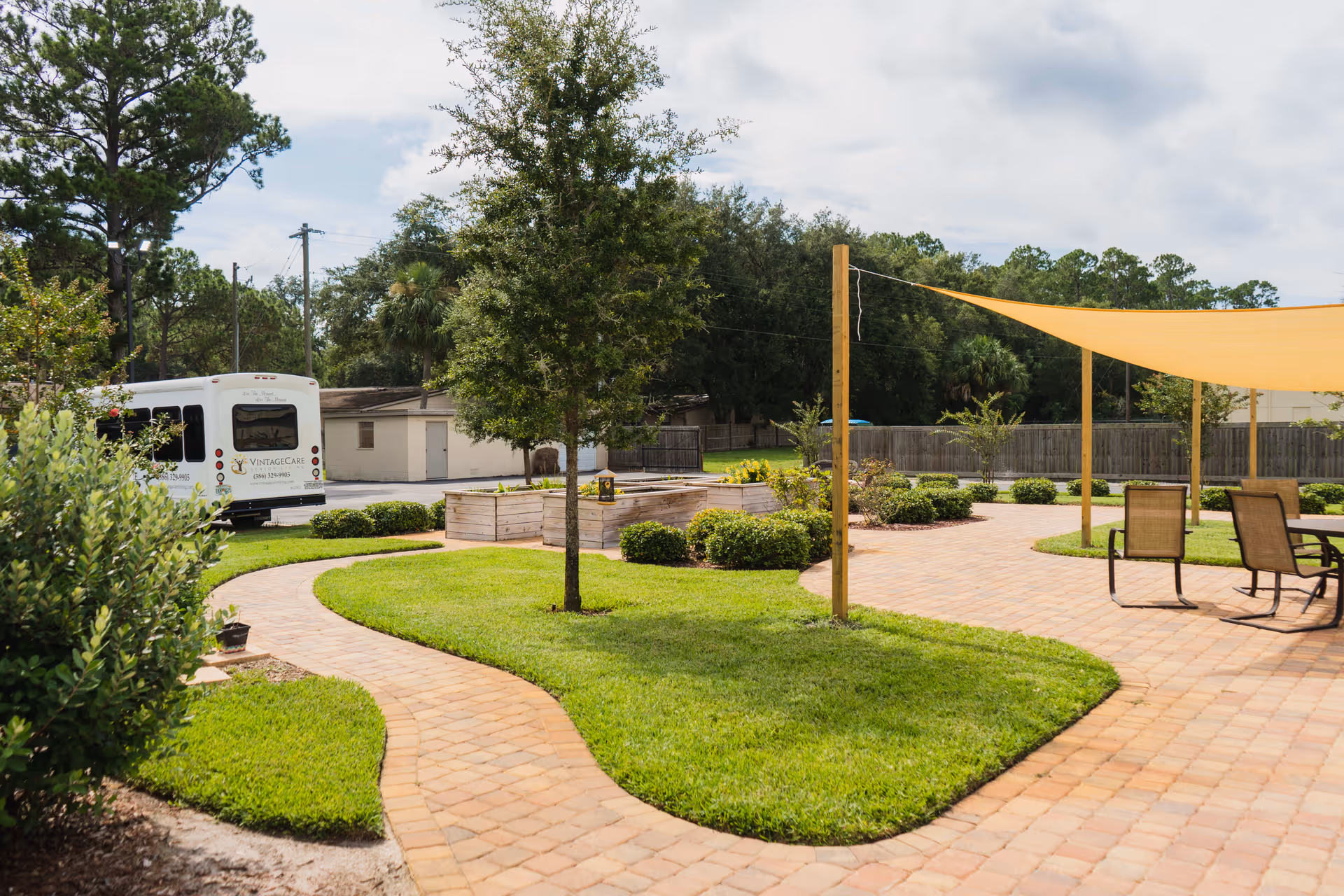 Outdoor garden area with a curved brick pathway, green grass, small trees and bushes, raised wooden garden beds, and a shaded seating area with chairs and a table under a yellow canopy. A white Vintage Care shuttle bus is parked near a small building in the background.