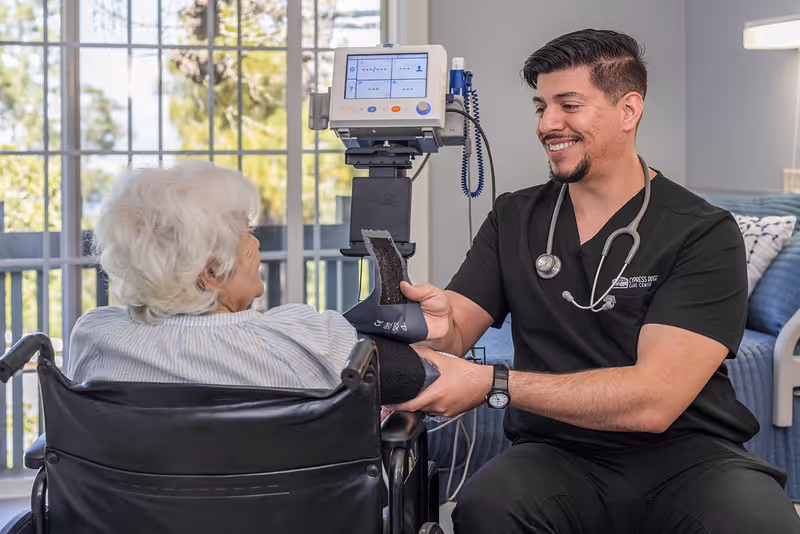 A healthcare professional wearing black scrubs and a stethoscope is smiling while taking the blood pressure of an elderly woman seated in a wheelchair near a large window with a view of trees outside.