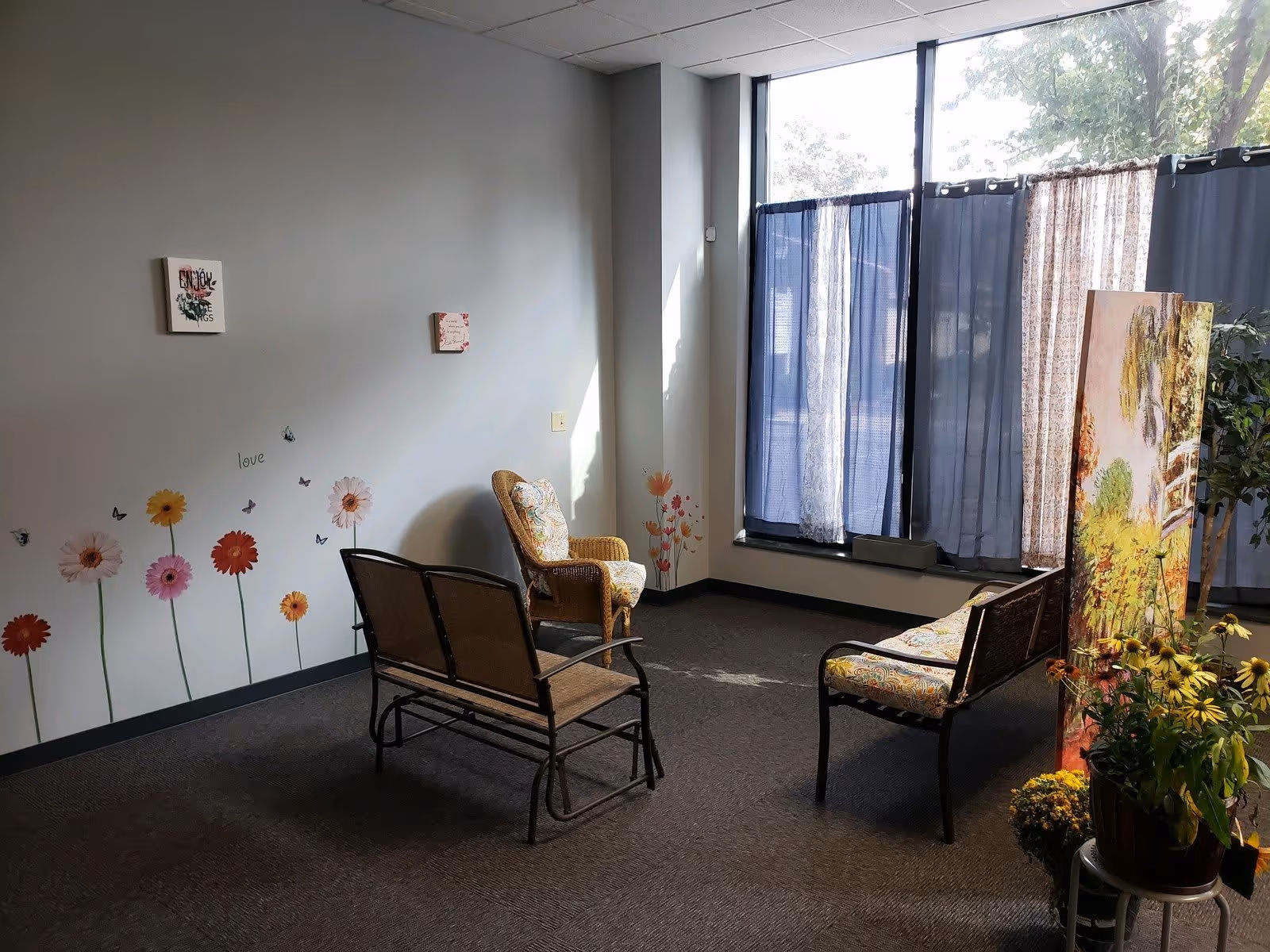 Sunlit sitting area with wicker and metal chairs, floral wall decals, potted plants, and large curtained windows.