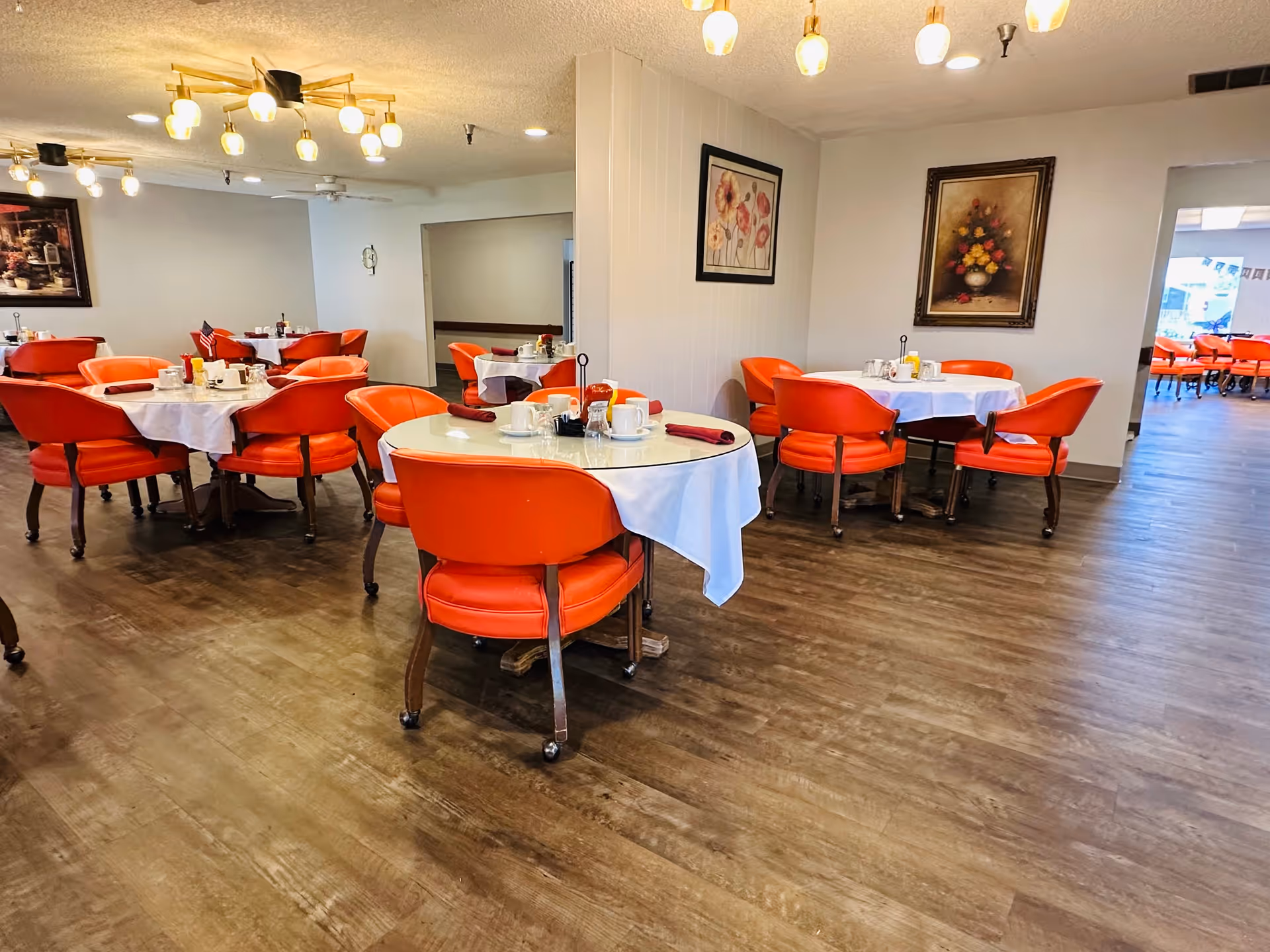 A senior living facility dining room with round tables covered in white tablecloths and set with cups, saucers, and condiments. The chairs are orange with wooden legs and wheels. The room has wooden flooring, framed floral paintings on the walls, and modern ceiling light fixtures.