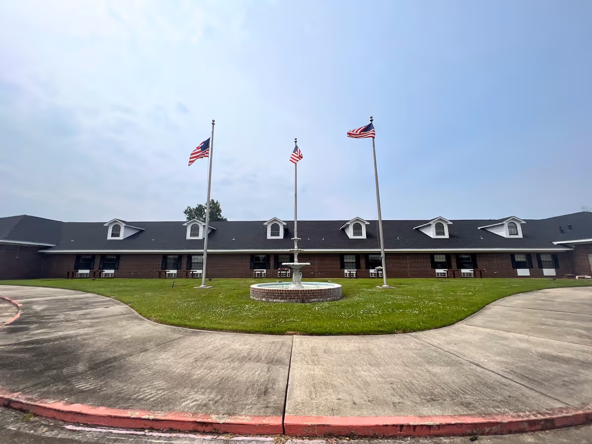 Front exterior view of a single-story brick building with a dark roof, featuring multiple dormer windows. In front of the building is a circular driveway surrounding a grassy area with a central fountain and three flagpoles flying American flags under a partly cloudy sky.