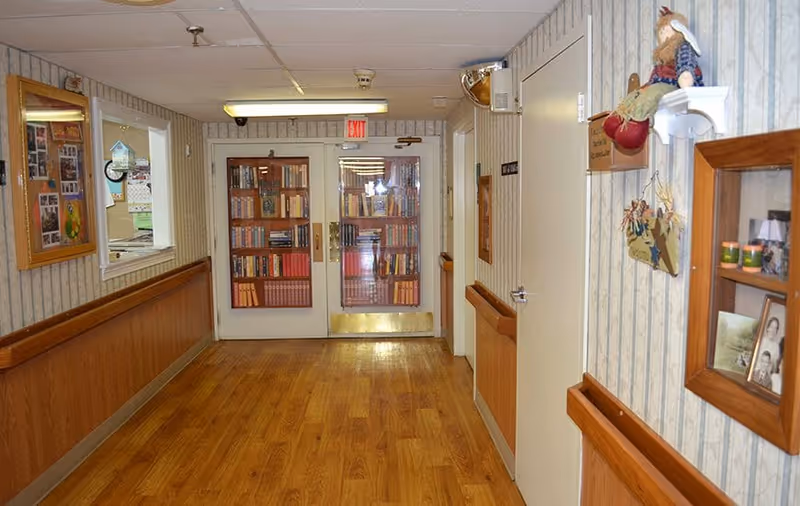 A hallway with wooden flooring and wallpapered walls featuring framed photos and decorations. At the end of the hallway, there are double doors with glass panels displaying bookshelves filled with books. The hallway has handrails on both sides and an exit sign above the doors.