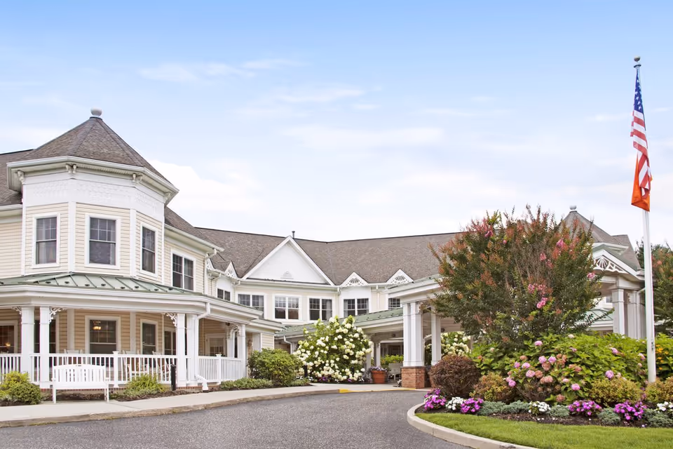 Exterior view of a senior living facility with a large, light-colored building featuring multiple windows, a covered entrance, and a landscaped garden with flowering bushes and an American flag on a flagpole.