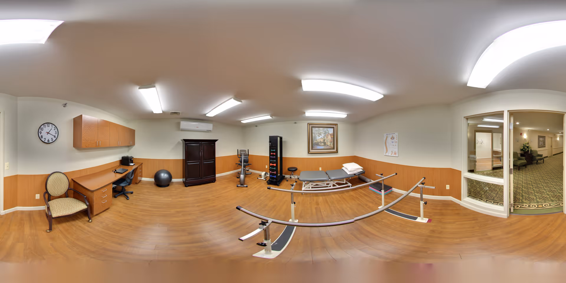 A spacious physical therapy or rehabilitation room with wooden flooring and white walls. The room contains parallel bars for walking exercises, a treatment table with pillows, a stationary exercise bike, a rack with colorful weights, an exercise ball, a desk with a chair, and a wall clock. There is a large window and door opening to a carpeted hallway with seating and plants.