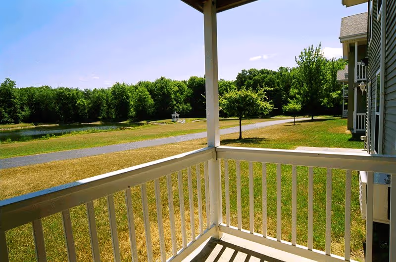 View from a covered balcony overlooking a grassy area with a paved walking path, a small pond, and trees in the background under a clear blue sky. Part of the building's exterior and balcony railing are visible.