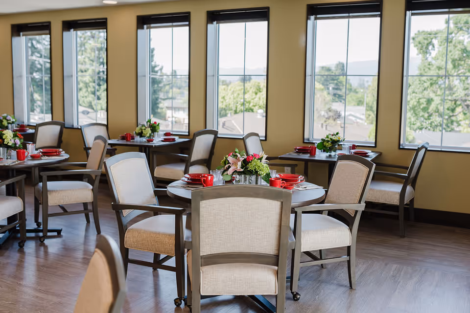 A bright dining room with multiple round tables set with red cups, plates, and silverware. Each table has a floral centerpiece. The room has large windows letting in natural light and offering a view of trees and rooftops outside. The chairs have light beige upholstery and wooden frames.