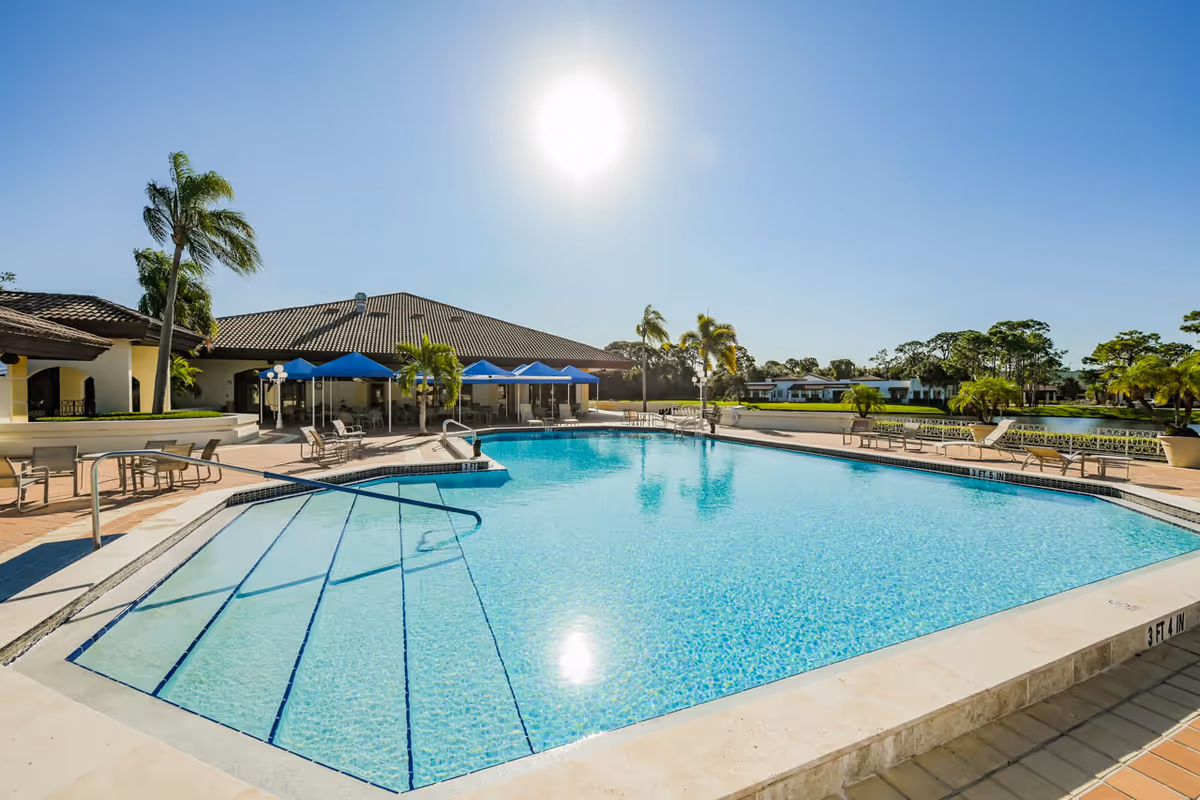 Outdoor swimming pool with clear blue water, surrounded by lounge chairs and tables with blue umbrellas. Palm trees and a building with a tiled roof are visible in the background under a bright sunny sky.