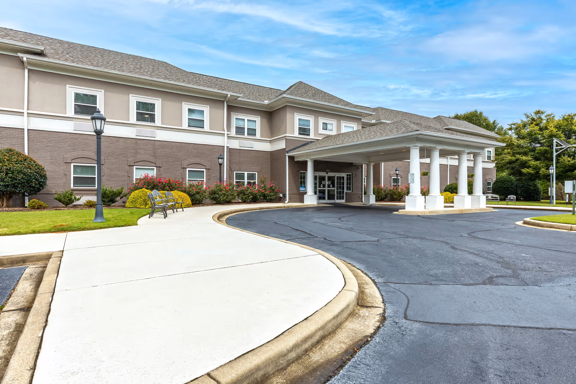 Exterior view of The Inverness at Spartanburg facility showing the main entrance with a covered drop-off area supported by white columns. The building has two stories with multiple windows, surrounded by landscaped bushes and flowers. There are benches and lamp posts along the sidewalk leading to the entrance under a partly cloudy blue sky.