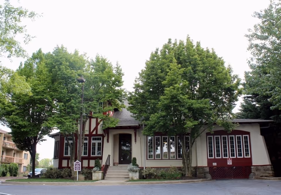 Exterior view of a senior living facility building with a beige and red facade, surrounded by green trees and shrubs, with a paved driveway in front.