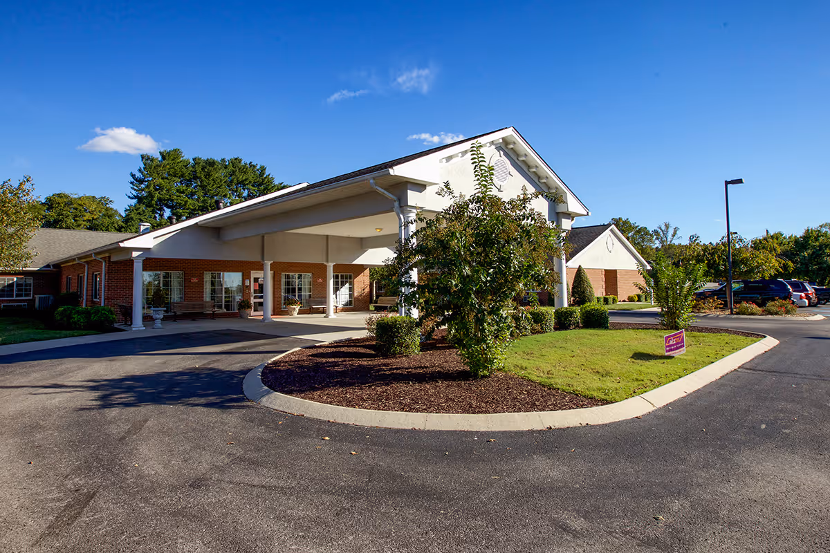 Exterior view of a single-story brick building with a covered entrance and a circular driveway, surrounded by green shrubs and trees under a clear blue sky.