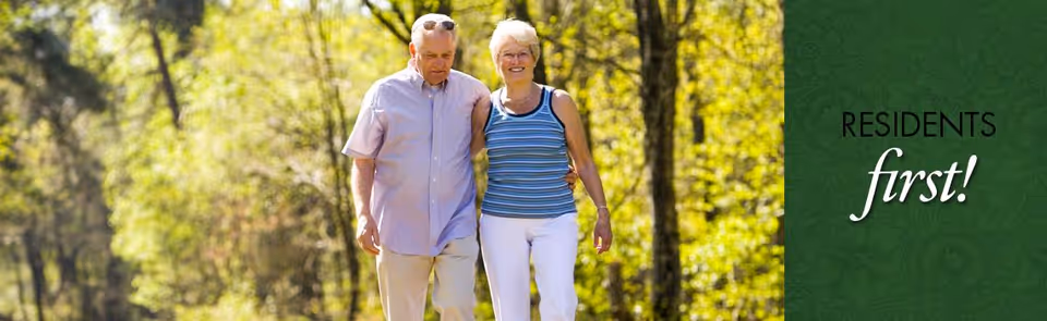 An elderly couple walking arm in arm along a sunlit wooded path, smiling and enjoying the outdoors.