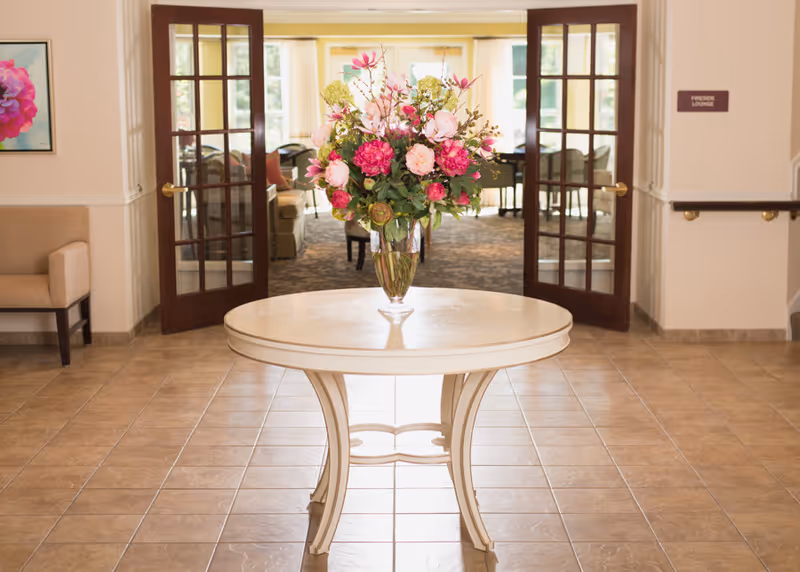 A round white table with a large vase of pink and white flowers is centered in a tiled hallway. Behind the table, double wooden doors with glass panes open into a lounge area with chairs and a piano. A beige armchair and a framed flower picture are visible on the left wall. A sign on the right wall reads 'Fireside Lounge'.