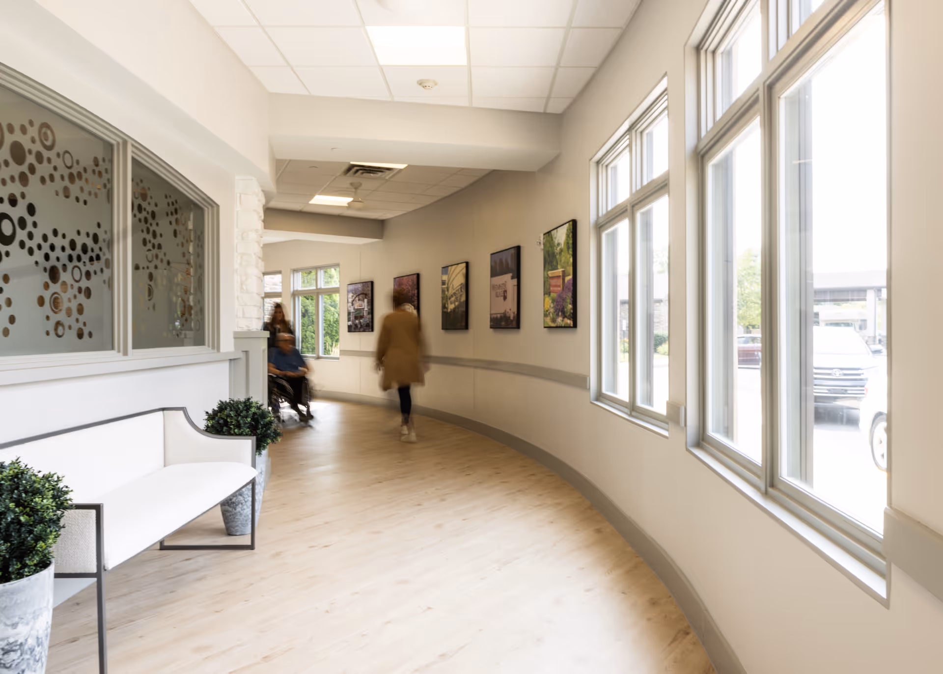 A bright, curved hallway in a senior living facility with large windows on the right side letting in natural light. On the left, there is a white bench with two potted plants beside it. The wall opposite the windows features framed pictures. Two people are visible in the hallway, one pushing another in a wheelchair.