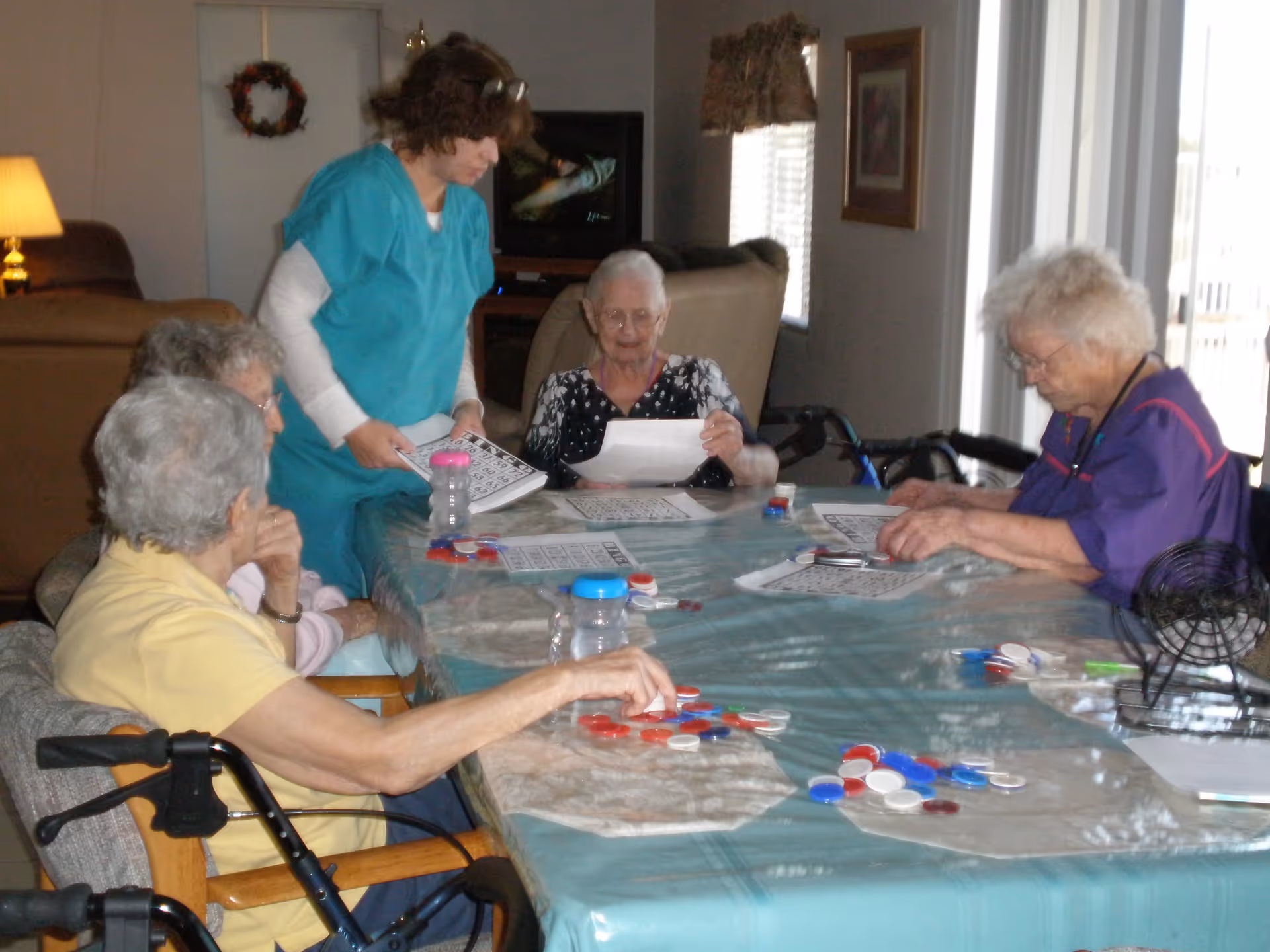 A group of elderly women sitting around a table playing bingo with a caregiver standing and assisting them. The table is covered with a blue tablecloth and bingo cards and chips are spread out. The room has a cozy interior with a lamp, a wreath on the wall, and a television in the background.