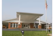 Front exterior view of a single-story building with a large covered entrance supported by white pillars, brick walls, multiple windows, and an American flag on a flagpole in front of the building.