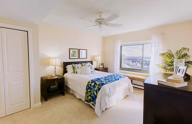 A bright and cozy bedroom with a neatly made bed featuring white bedding and a blue floral throw. There are two bedside tables with lamps, framed botanical prints on the wall above the bed, a large window with a view, a cushioned bench beneath the window, a dark wooden dresser with books and a plant, and light-colored carpeted flooring.