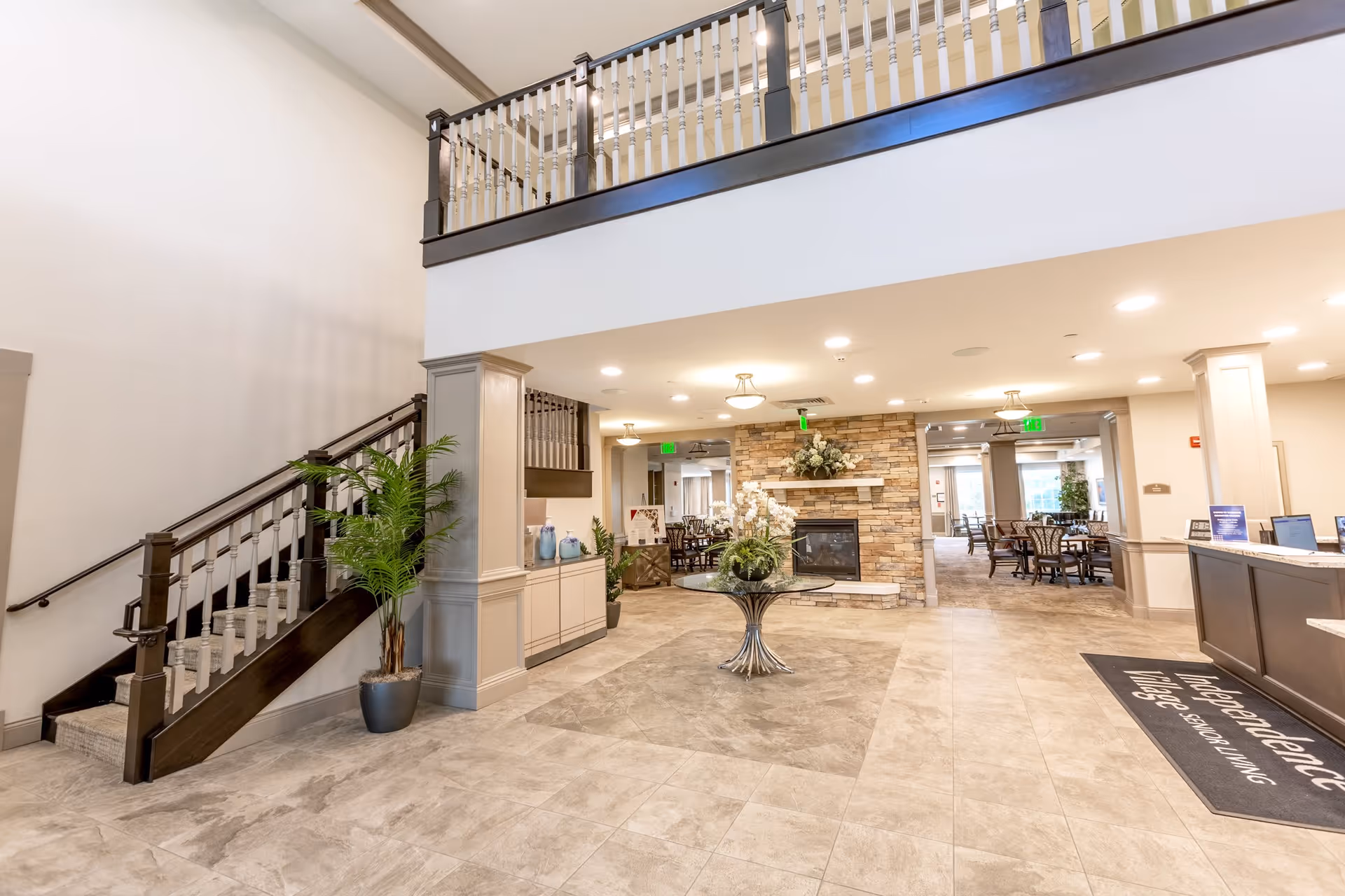 Spacious and well-lit interior lobby area of Independence Village of Zionsville West featuring a staircase with dark wood railing on the left, a round glass table with a floral arrangement in the center, a stone fireplace with decorative flowers, and a reception desk on the right with a mat displaying the facility's name.