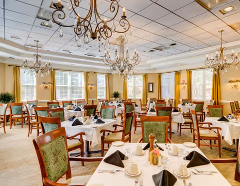 A bright and elegant dining room with multiple tables covered in white tablecloths, set with cups, plates, silverware, and black folded napkins. The room features green upholstered wooden chairs, large windows with yellow curtains, and ornate chandeliers hanging from the ceiling.