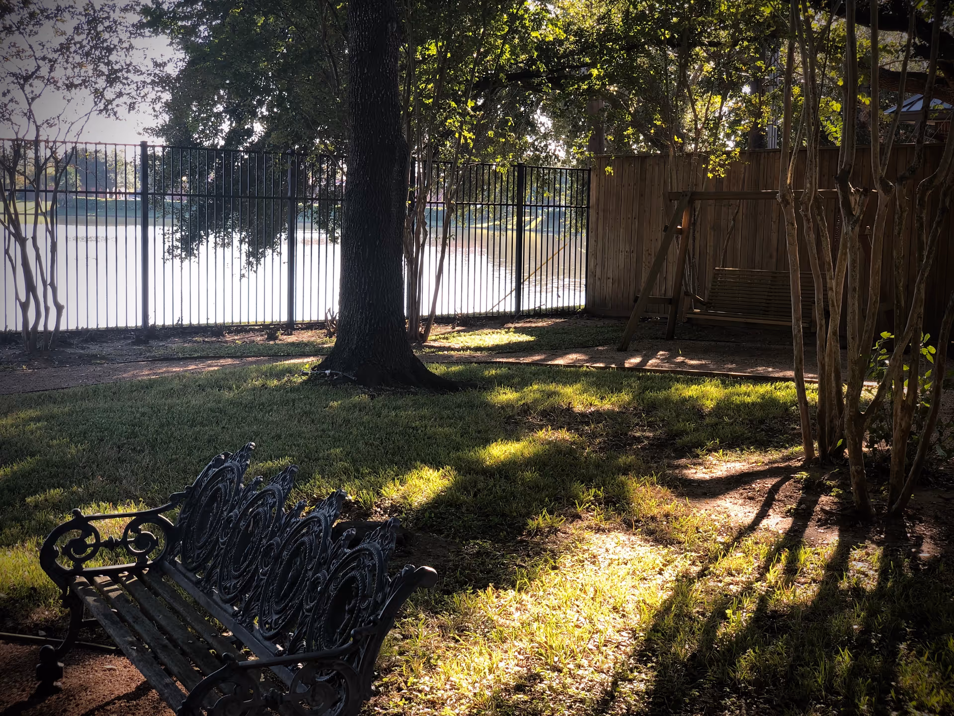 A shaded outdoor garden area with a decorative metal bench in the foreground and a wooden swing bench in the background. The area is surrounded by trees and a metal fence, with sunlight filtering through the leaves and casting shadows on the grass.
