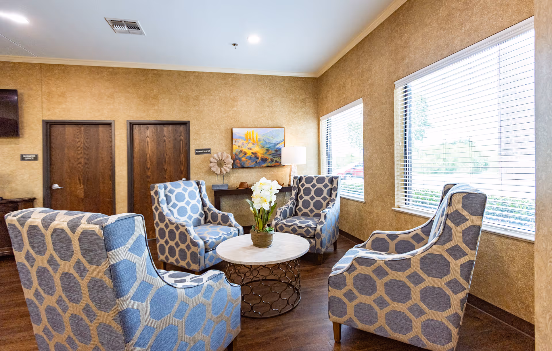 A cozy sitting area in a senior living facility with four patterned armchairs arranged around a round coffee table with a potted white orchid. The room has beige textured walls, two large windows with blinds, wooden doors labeled 'Conference' and 'Administration,' a small console table with decorative items, and a wall-mounted TV.