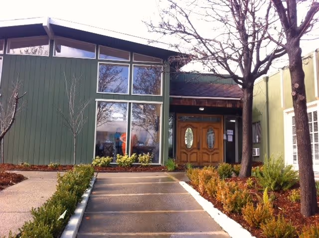 Exterior view of a building with green siding and large windows reflecting trees. The entrance features double wooden doors with oval glass panels, surrounded by landscaped bushes and trees along a paved walkway.