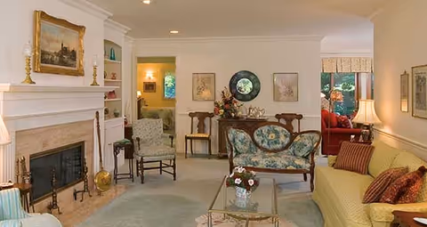A cozy living room with a fireplace on the left, a glass coffee table with a flower arrangement in the center, a floral upholstered loveseat, a yellow sofa with red and brown pillows, and various chairs. The room has soft lighting, framed artwork on the walls, and an adjacent room with red furniture visible through an open doorway.