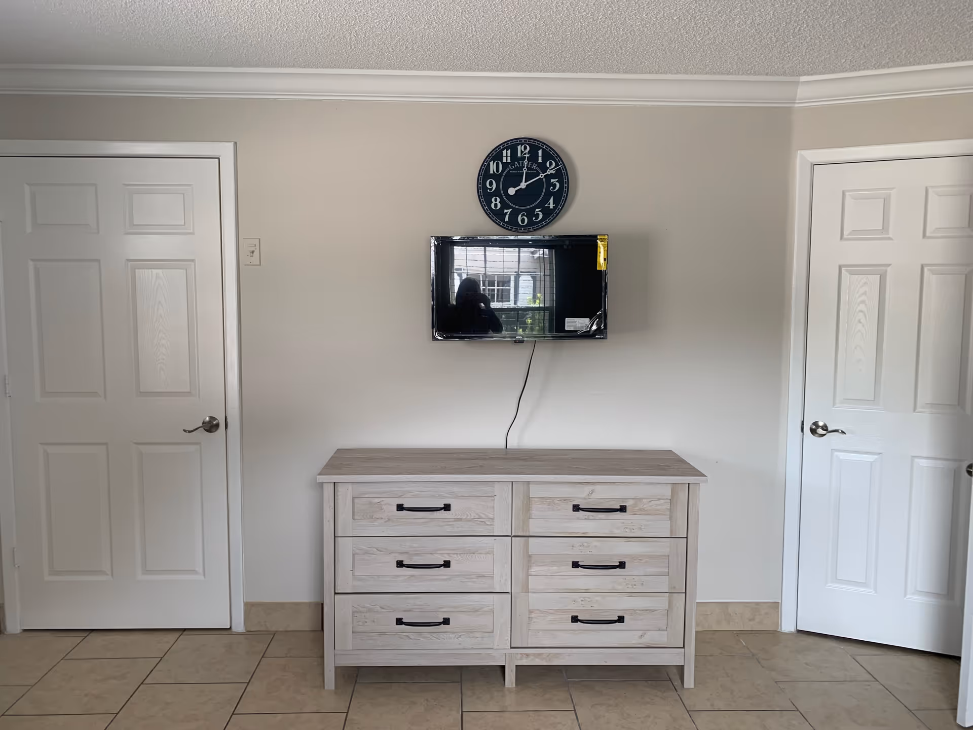 A simple interior wall with two white doors on either side, a wooden dresser with six drawers in the center, a flat-screen TV mounted above the dresser, and a round black wall clock above the TV showing the time as 11:55.