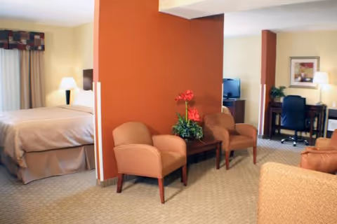 A cozy interior space featuring a bedroom area with a neatly made bed and bedside lamp, separated by a partial wall from a sitting area with two armchairs and a small table with a flower arrangement. In the background, there is a desk with a chair, a lamp, and a television on a stand.
