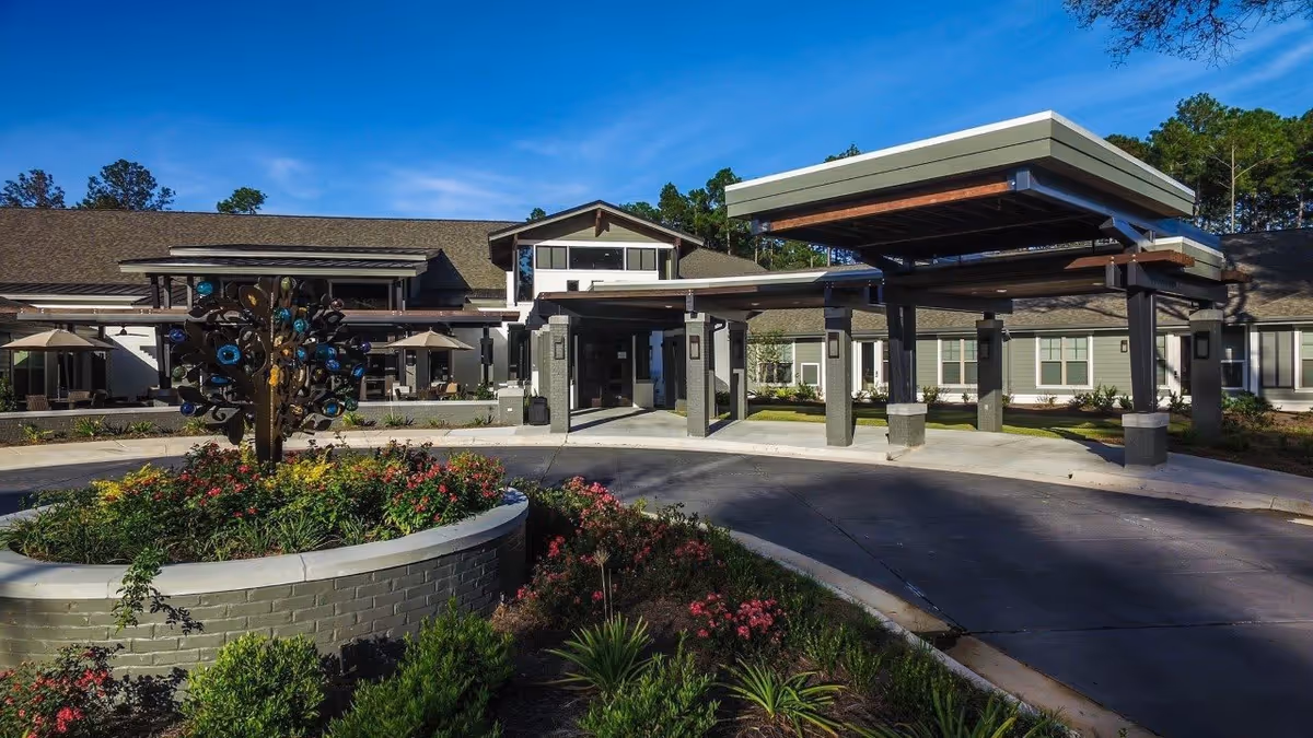 Exterior view of Avanti Senior Living at Covington showing the main entrance with a covered drop-off area, surrounded by landscaped gardens with flowers and a decorative metal sculpture in the center of a circular planter.